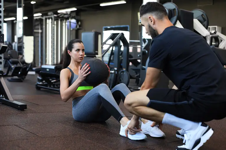 Trainer helping young woman to do abdominal exercises in gym.